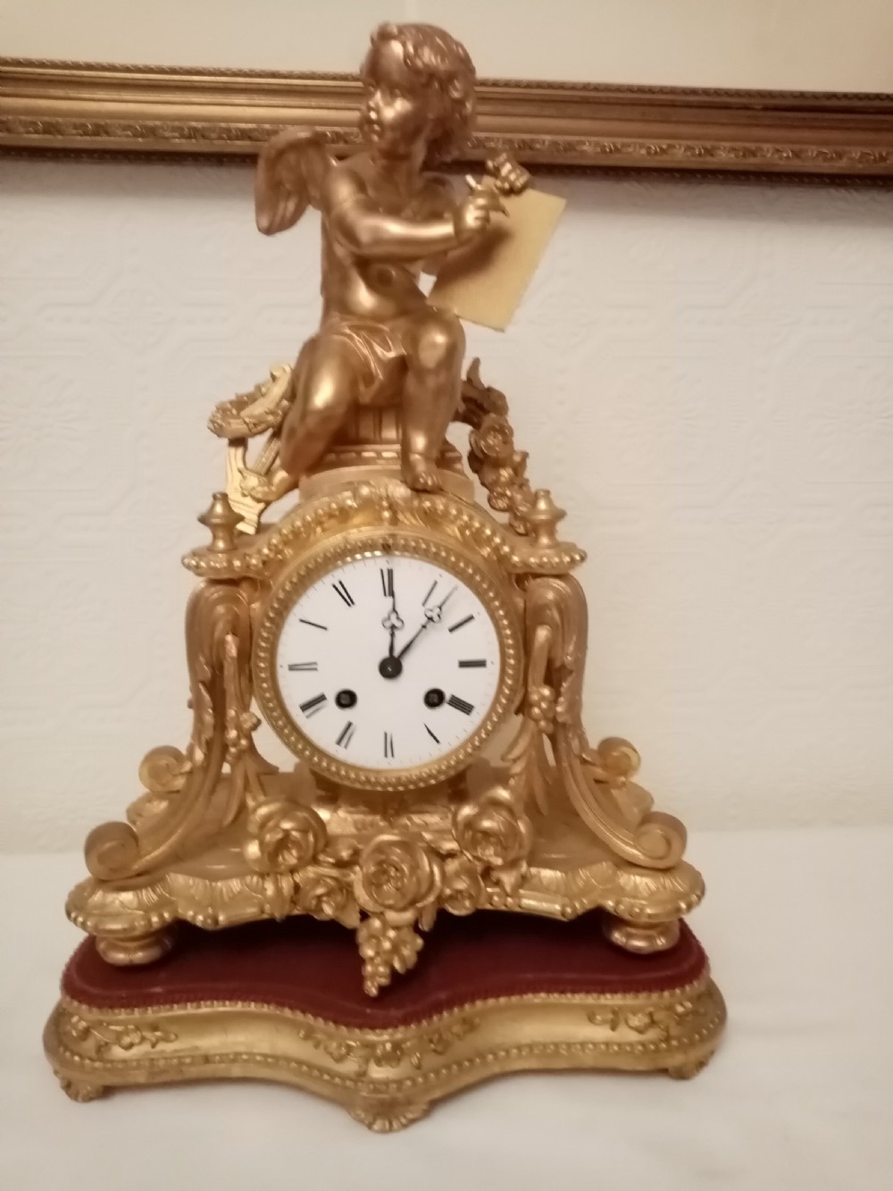 An ornate gold gilt bronze mantel clock topped with a seated cherub figure writing on a scroll above an enamelled dial with Roman numerals.
