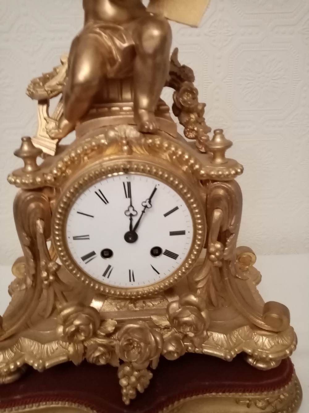 An ornate gold gilt bronze mantel clock topped with a seated cherub figure writing on a scroll above an enamelled dial with Roman numerals. View III