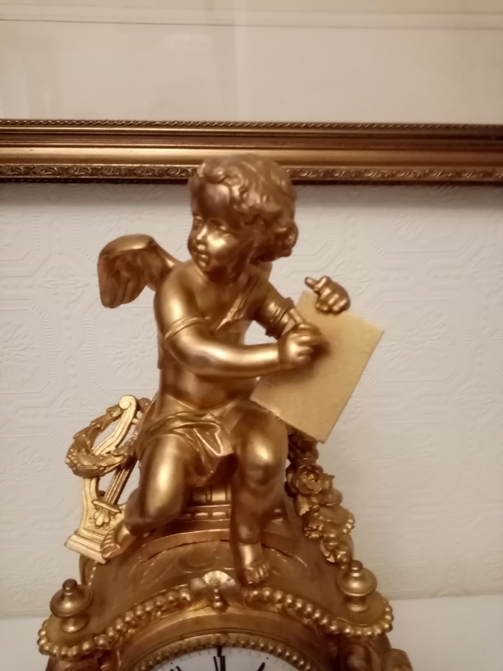 An ornate gold gilt bronze mantel clock topped with a seated cherub figure writing on a scroll above an enamelled dial with Roman numerals. View VI
