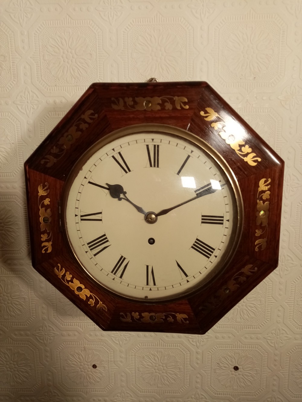 An octagonal dark wood wall clock featuring brass foliate inlay surrounding a cream dial with Roman numerals and black hands.