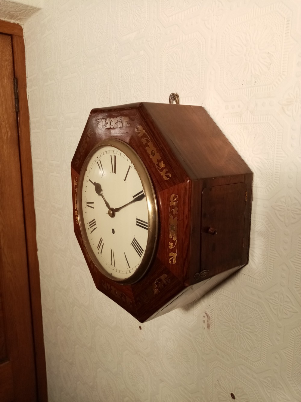 An octagonal dark wood wall clock featuring brass foliate inlay surrounding a cream dial with Roman numerals and black hands. View II