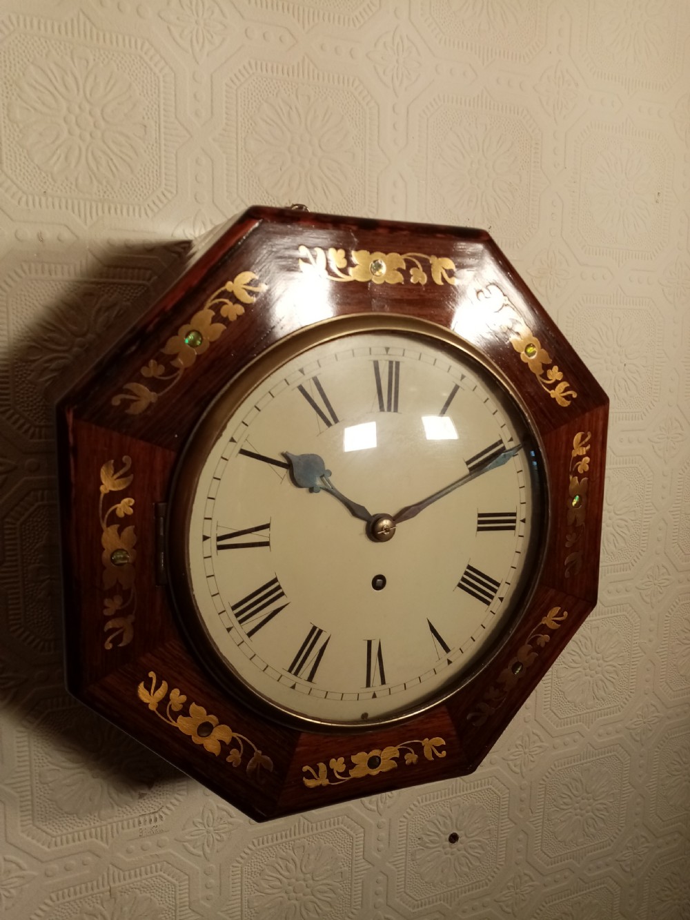 An octagonal dark wood wall clock featuring brass foliate inlay surrounding a cream dial with Roman numerals and black hands. View III
