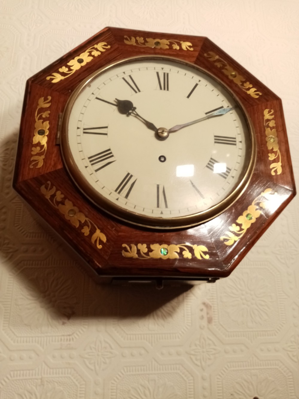 An octagonal dark wood wall clock featuring brass foliate inlay surrounding a cream dial with Roman numerals and black hands. View IV