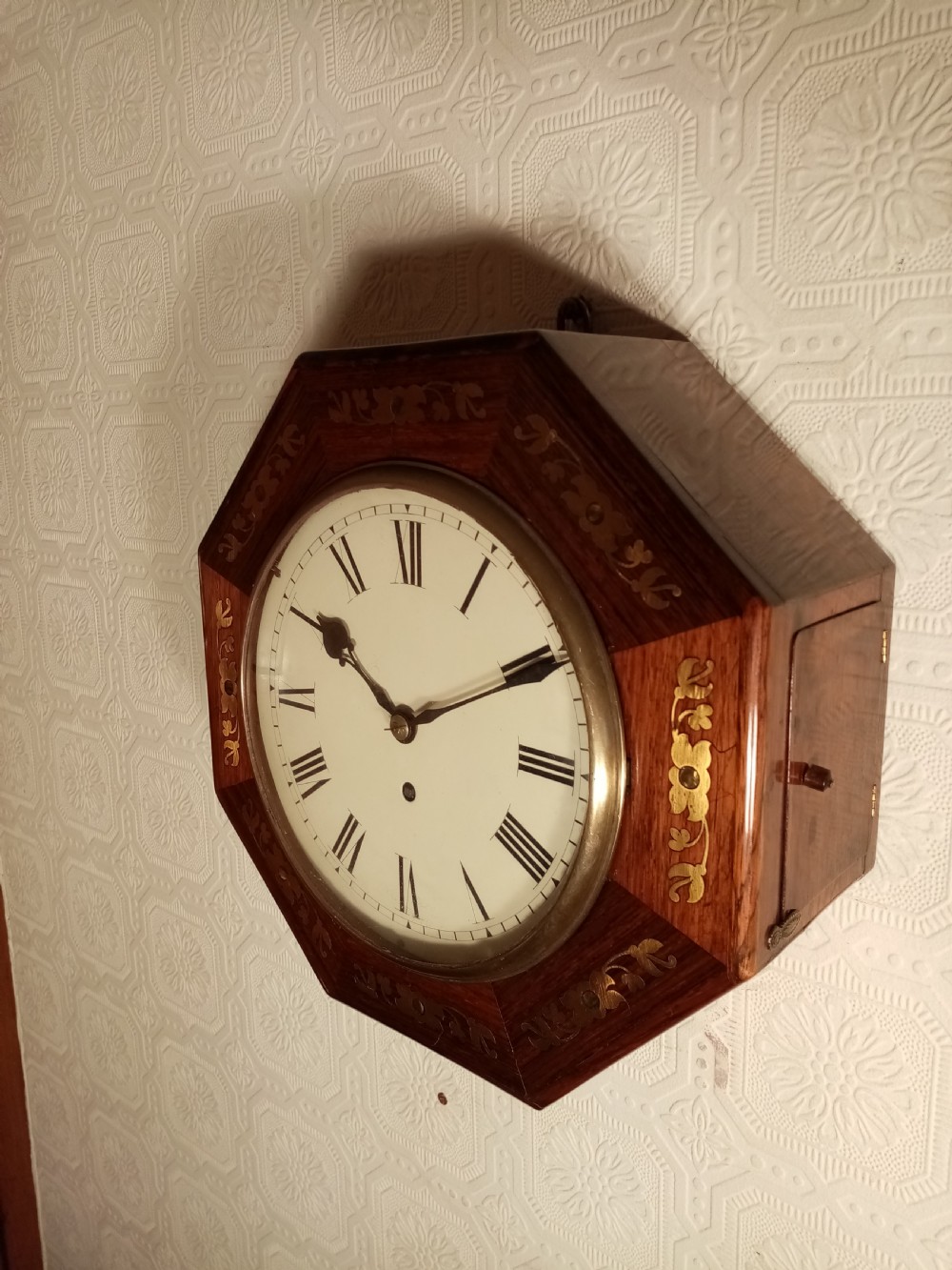 An octagonal dark wood wall clock featuring brass foliate inlay surrounding a cream dial with Roman numerals and black hands. View V