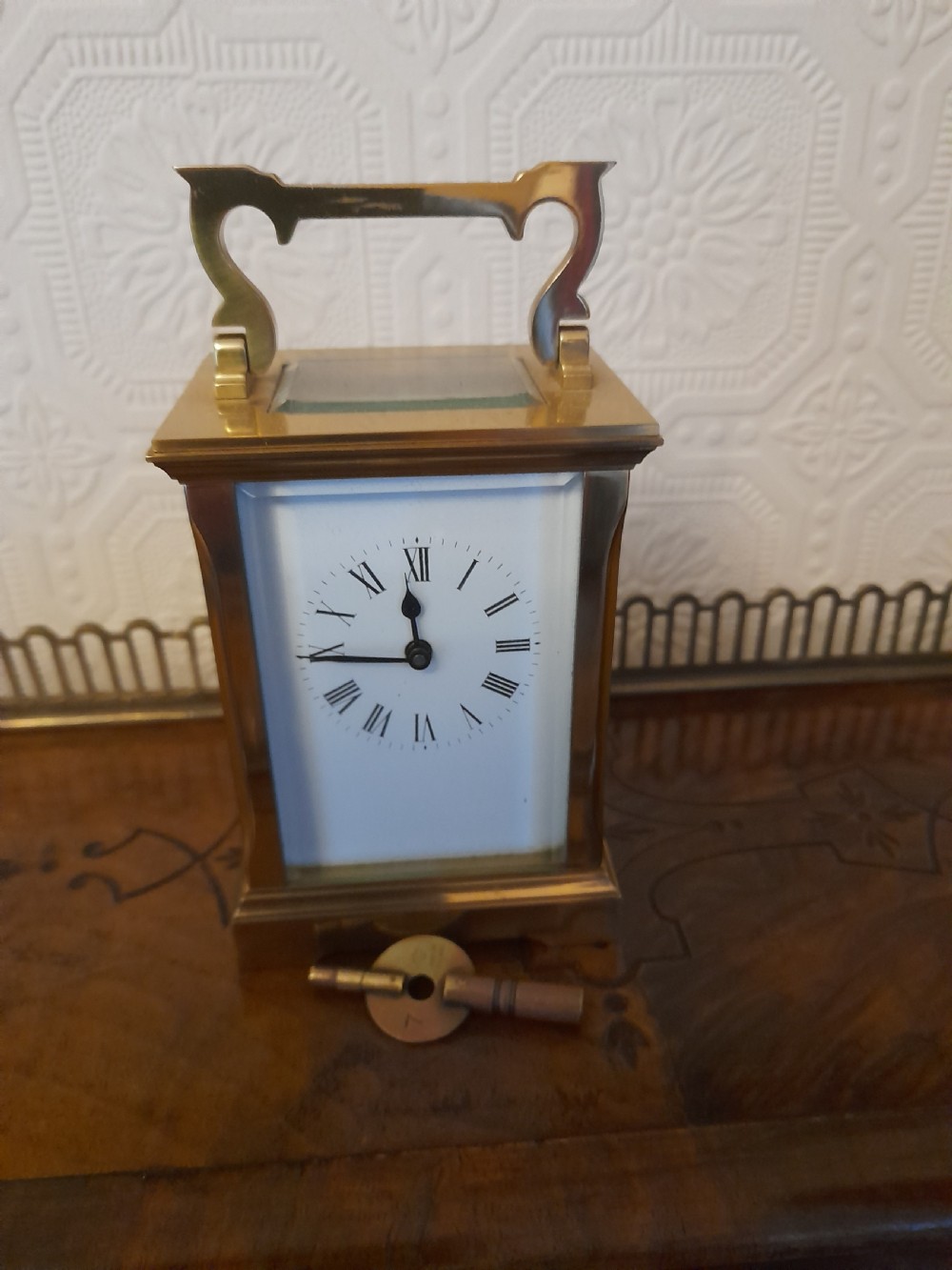 A rectangular brass carriage clock featuring clear glass side panels, a white enamel dial with black Roman numerals, and a shaped brass carrying handle.