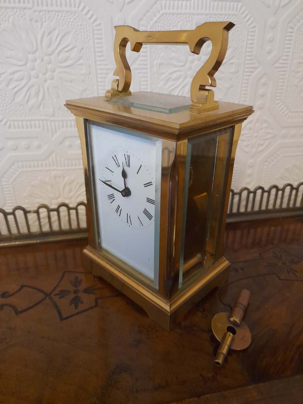 A rectangular brass carriage clock featuring clear glass side panels, a white enamel dial with black Roman numerals, and a shaped brass carrying handle. View II