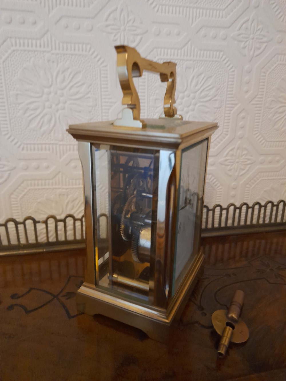A rectangular brass carriage clock featuring clear glass side panels, a white enamel dial with black Roman numerals, and a shaped brass carrying handle. View III