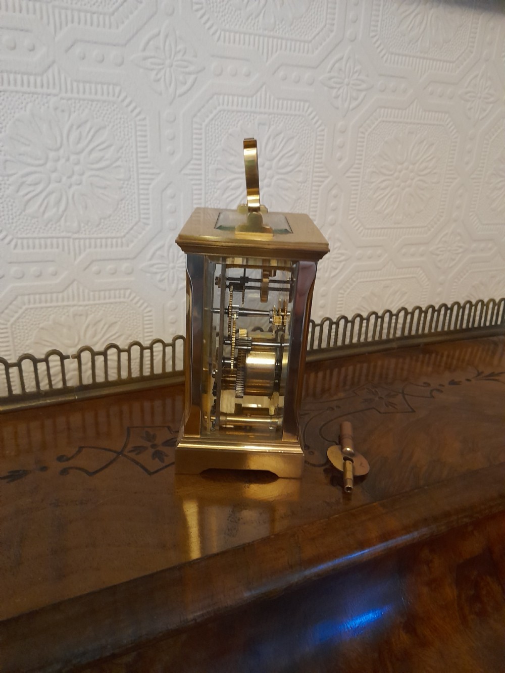 A rectangular brass carriage clock featuring clear glass side panels, a white enamel dial with black Roman numerals, and a shaped brass carrying handle. View VI