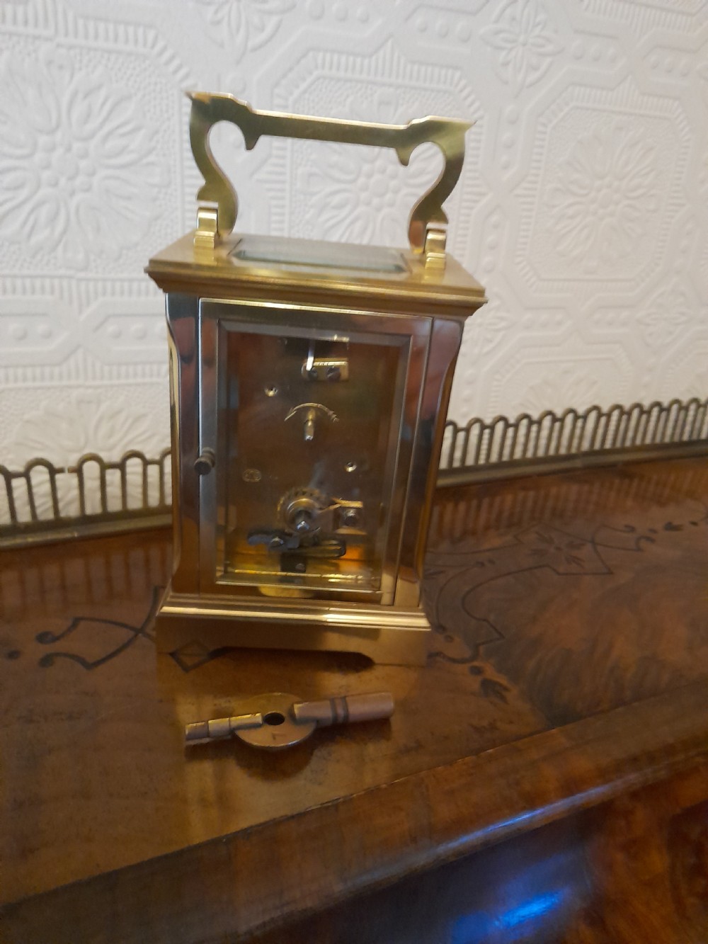 A rectangular brass carriage clock featuring clear glass side panels, a white enamel dial with black Roman numerals, and a shaped brass carrying handle. View VII