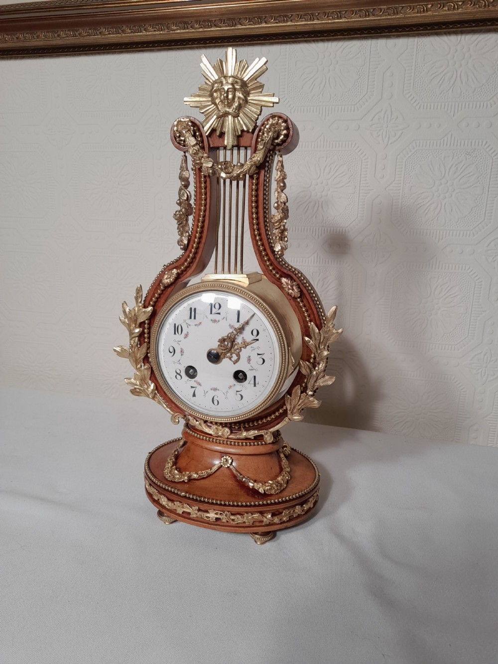 A Louis XVI style wooden lyre clock featuring gilt bronze floral mounts, a sunburst mask finial, and a white enamel dial.