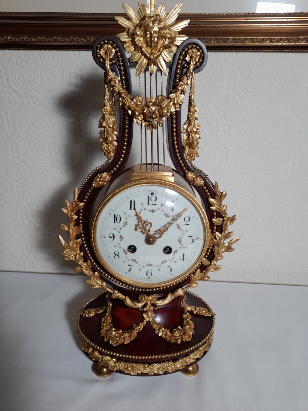 A Louis XVI style lyre-shaped mantel clock crafted from dark mahogany with elaborate gilt bronze floral festoons, an Apollo sunburst finial, and a floral enamel dial.