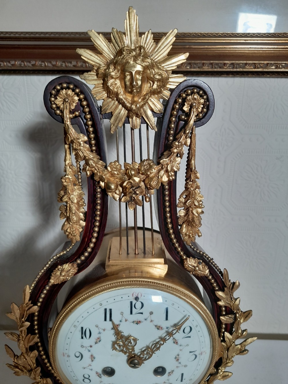 A Louis XVI style lyre-shaped mantel clock crafted from dark mahogany with elaborate gilt bronze floral festoons, an Apollo sunburst finial, and a floral enamel dial. View VII