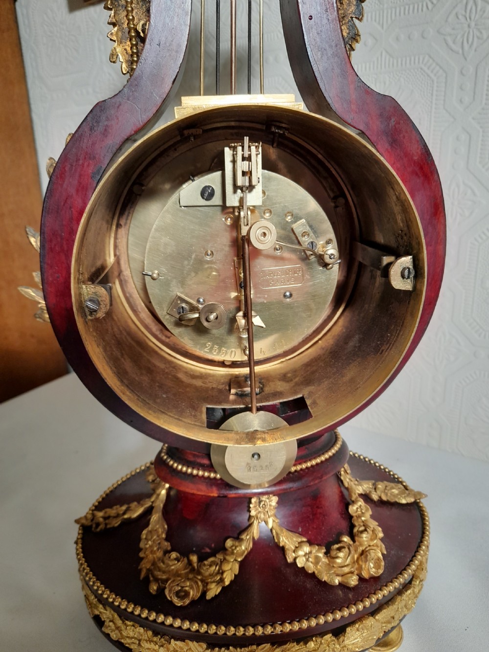 A Louis XVI style lyre-shaped mantel clock crafted from dark mahogany with elaborate gilt bronze floral festoons, an Apollo sunburst finial, and a floral enamel dial. View VIII
