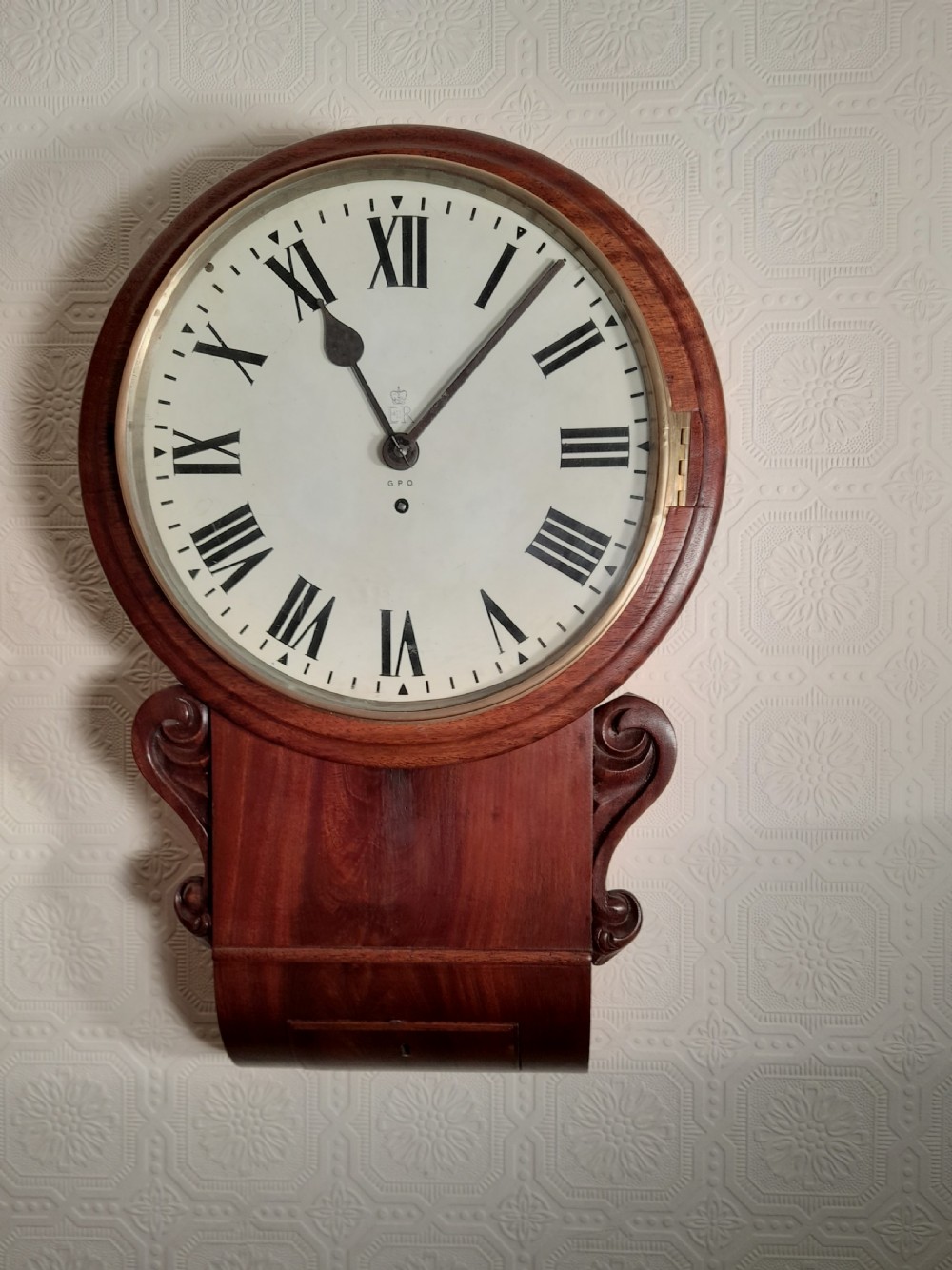 An antique mahogany drop dial wall clock featuring an off-white circular dial face with black Roman numerals, a brass bezel, and carved wooden side flourishes.