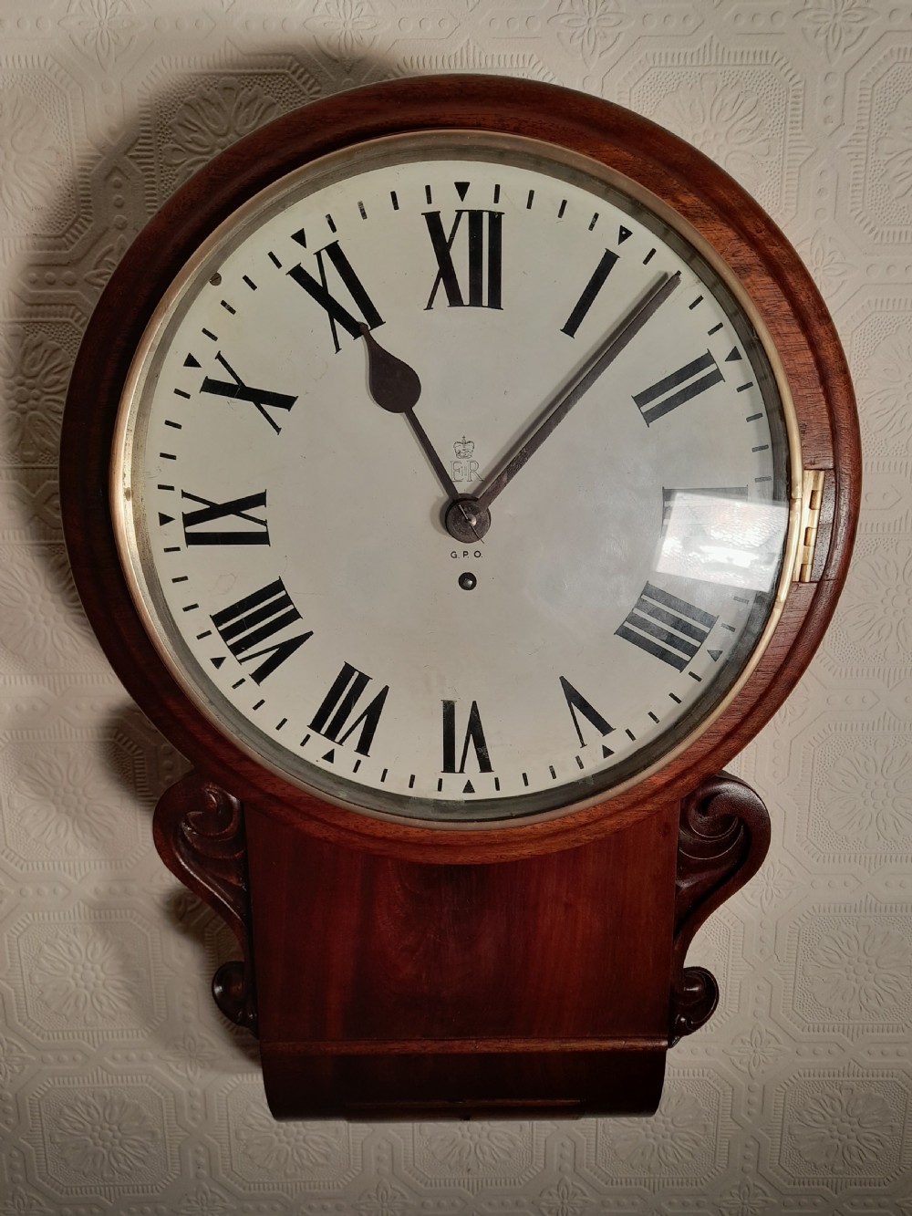 An antique mahogany drop dial wall clock featuring an off-white circular dial face with black Roman numerals, a brass bezel, and carved wooden side flourishes. View III