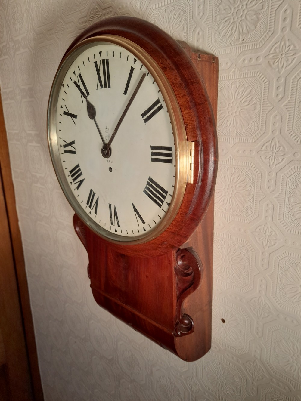 An antique mahogany drop dial wall clock featuring an off-white circular dial face with black Roman numerals, a brass bezel, and carved wooden side flourishes. View IV