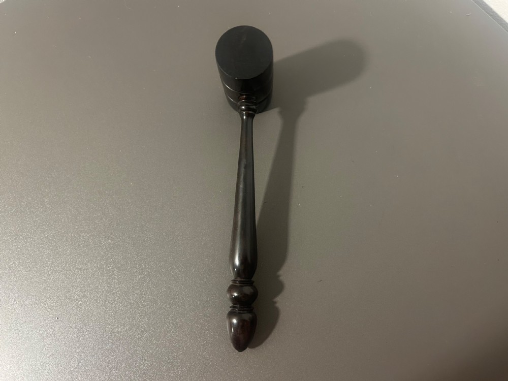 A turned dark ebony wood gavel featuring a cylindrical head with incised ring details and a lathe-turned handle ending in a rounded finial. View VI
