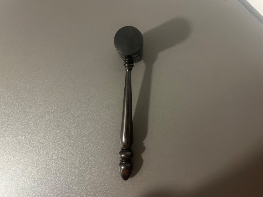 A turned dark ebony wood gavel featuring a cylindrical head with incised ring details and a lathe-turned handle ending in a rounded finial. View V