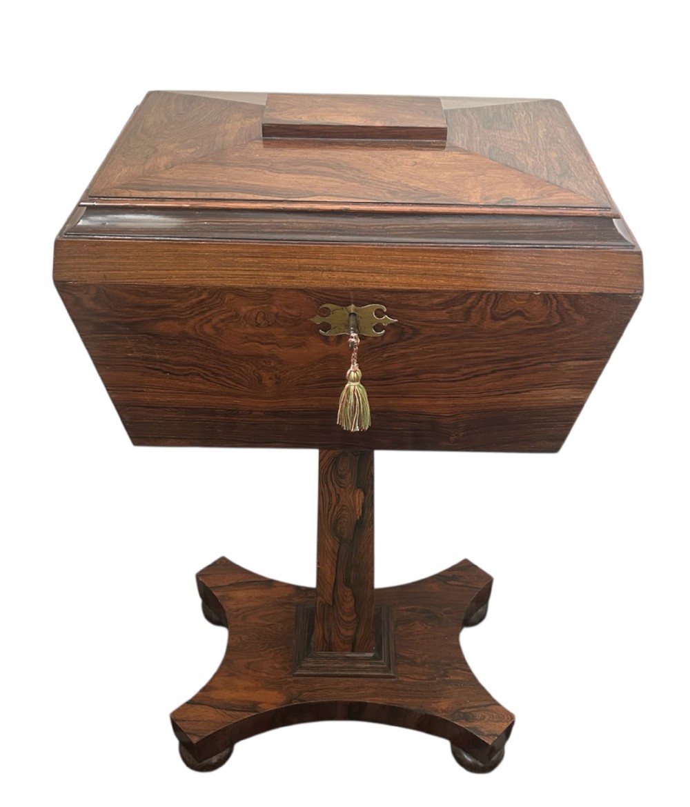 A Regency rosewood teapoy featuring a sarcophagus-shaped caddy top, brass escutcheon with key and tassel, set upon a central column and a four-pointed concave base.