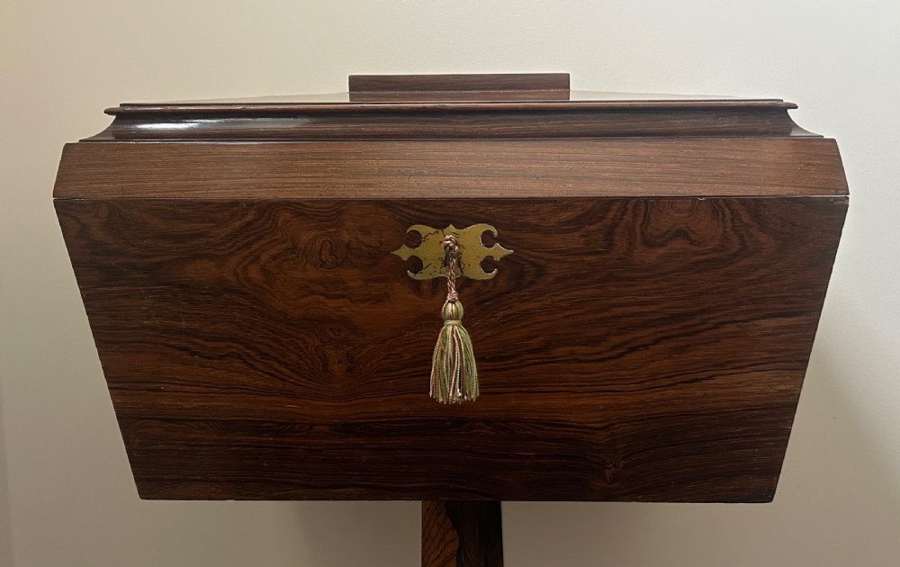 A Regency rosewood teapoy featuring a sarcophagus-shaped caddy top, brass escutcheon with key and tassel, set upon a central column and a four-pointed concave base. View VI