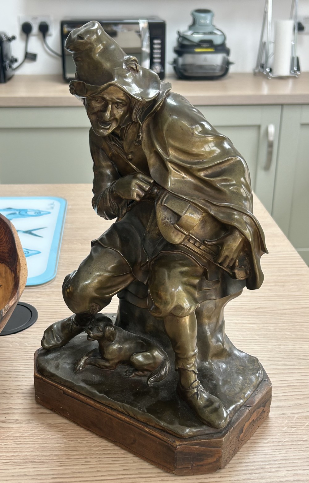 A patinated dark bronze sculpture depicting a seated male musician with a hurdy-gurdy instrument and a small dog at his feet on a wooden plinth. View II