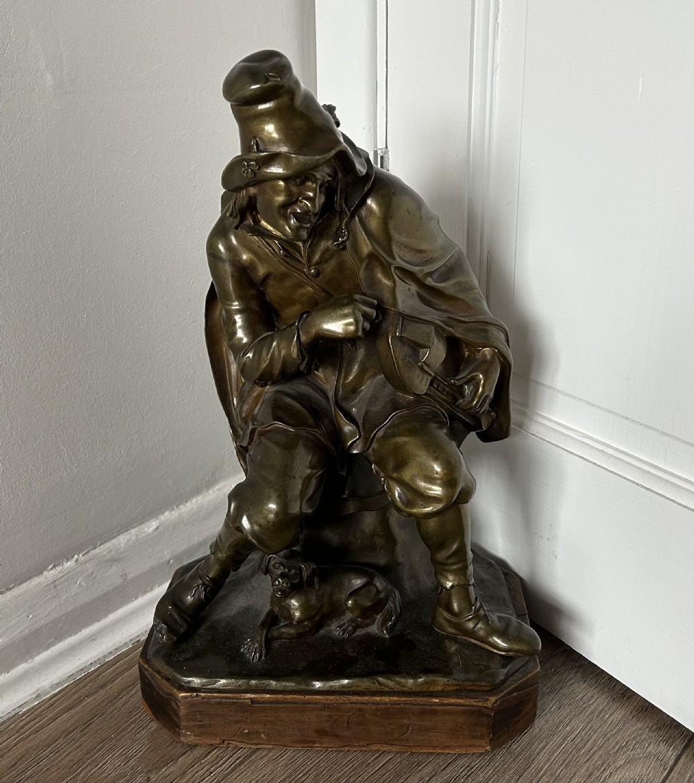 A patinated dark bronze sculpture depicting a seated male musician with a hurdy-gurdy instrument and a small dog at his feet on a wooden plinth. View IX