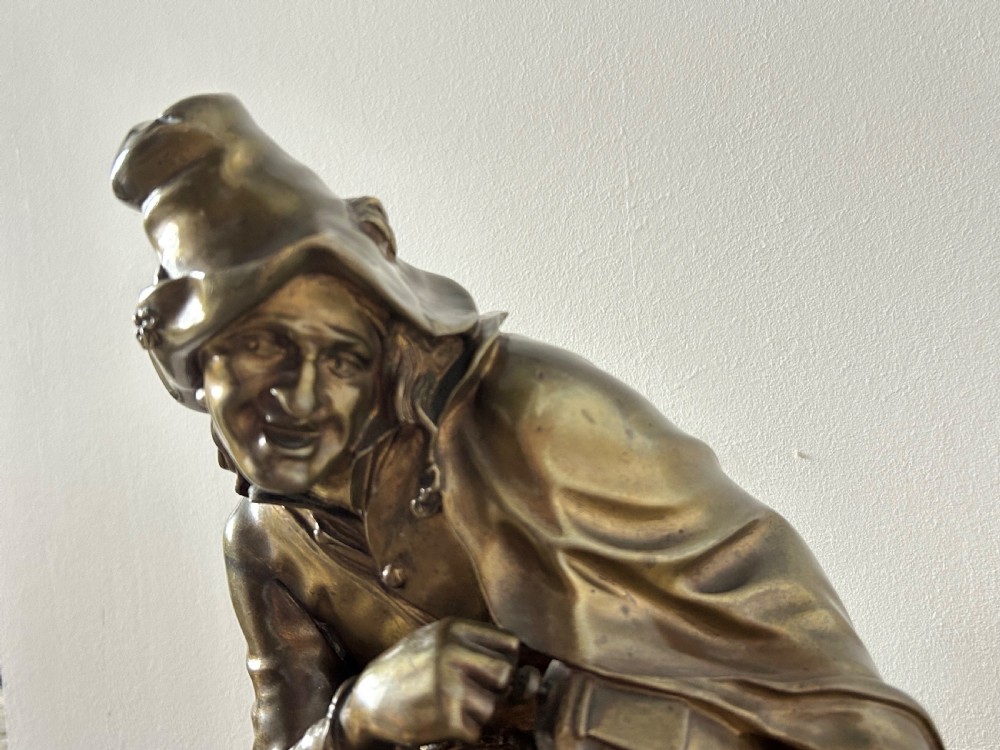A patinated dark bronze sculpture depicting a seated male musician with a hurdy-gurdy instrument and a small dog at his feet on a wooden plinth. View XI