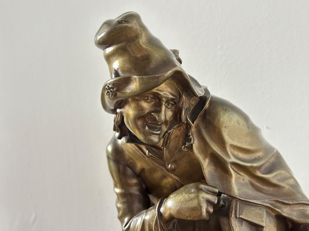 A patinated dark bronze sculpture depicting a seated male musician with a hurdy-gurdy instrument and a small dog at his feet on a wooden plinth. View XII