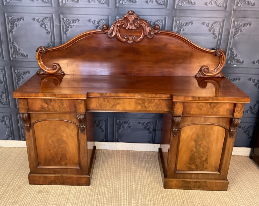 A Victorian mahogany pedestal sideboard featuring a carved arched backboard, flame mahogany veneer panels, and twin pedestal cupboards.