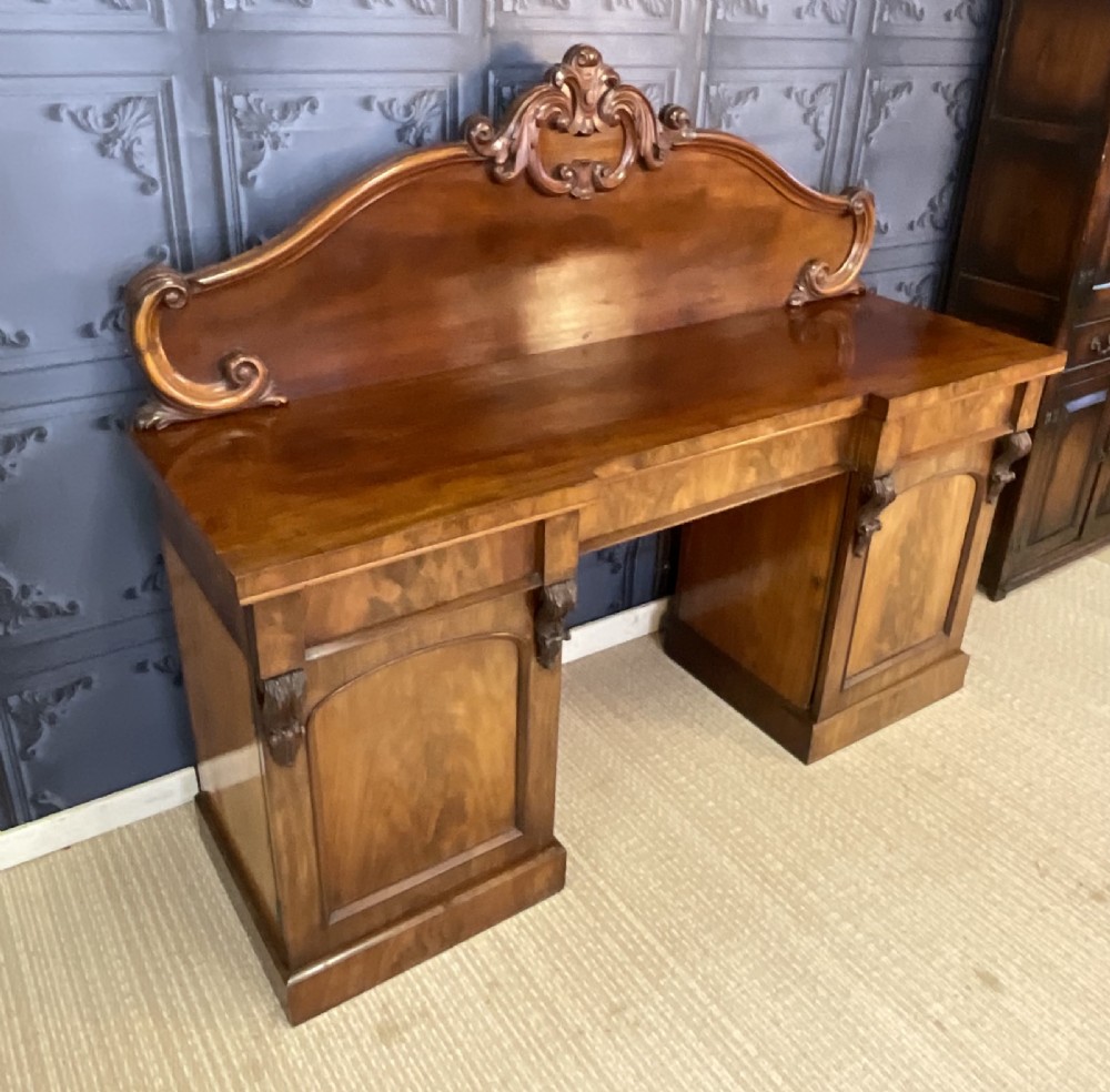 A Victorian mahogany pedestal sideboard featuring a carved arched backboard, flame mahogany veneer panels, and twin pedestal cupboards. View V