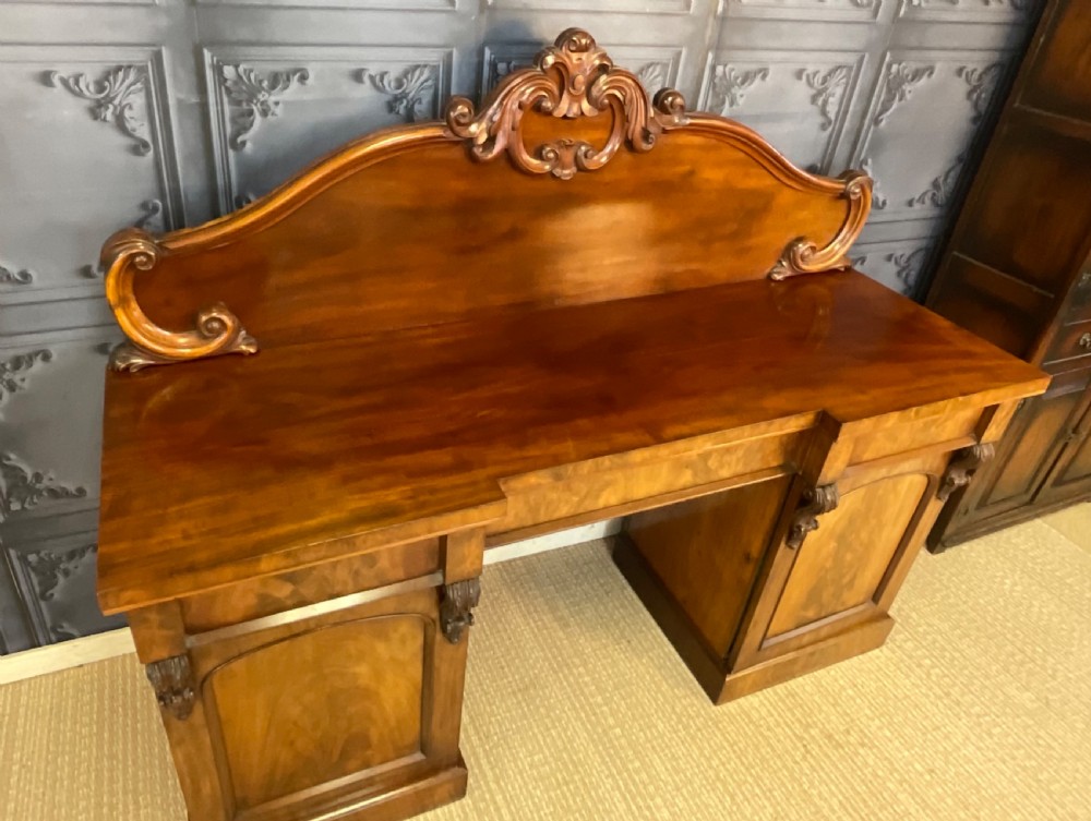A Victorian mahogany pedestal sideboard featuring a carved arched backboard, flame mahogany veneer panels, and twin pedestal cupboards. View VI