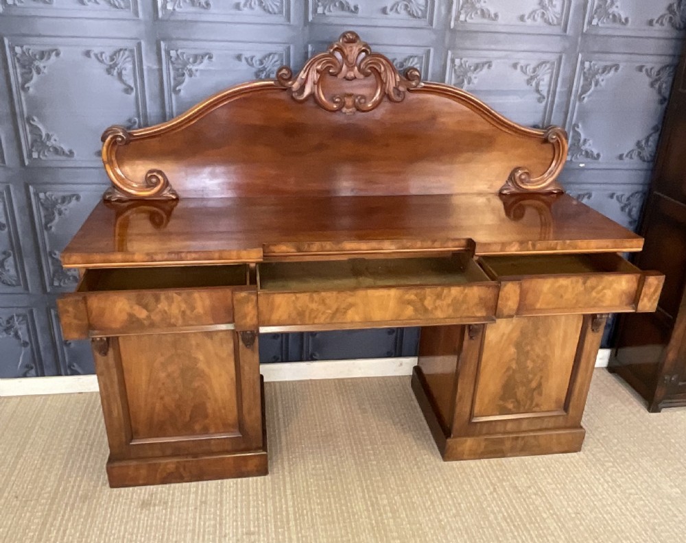 A Victorian mahogany pedestal sideboard featuring a carved arched backboard, flame mahogany veneer panels, and twin pedestal cupboards. View VII