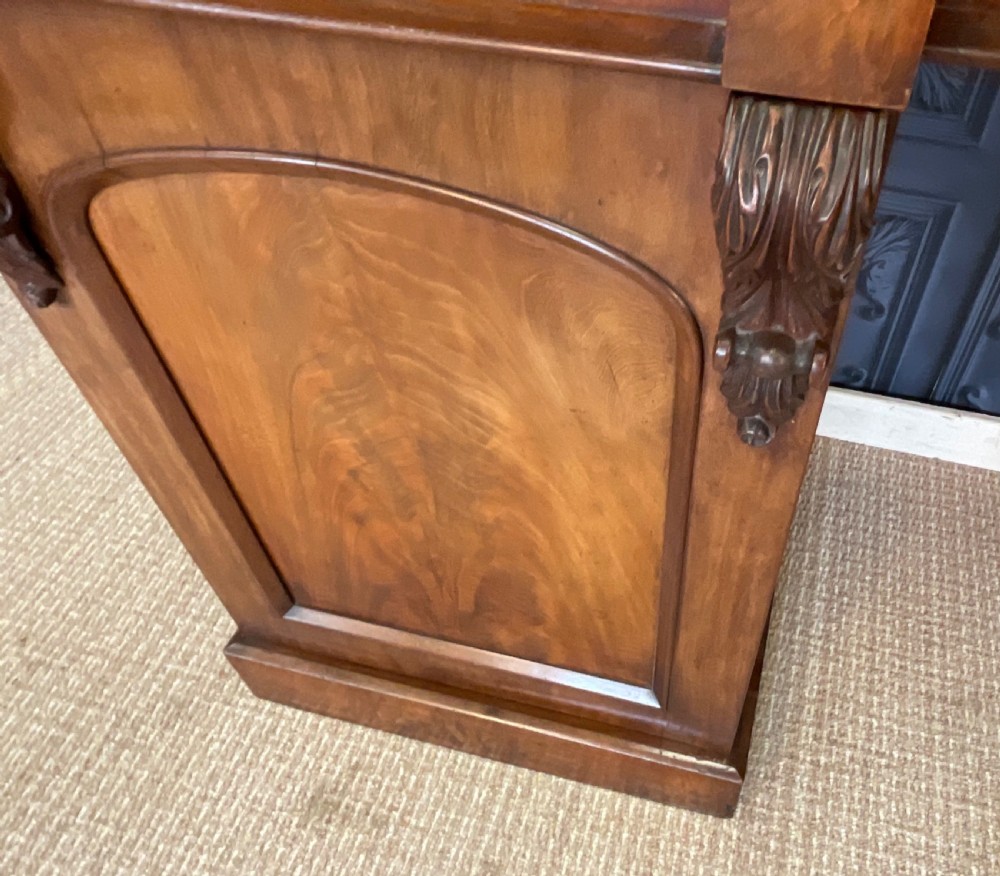 A Victorian mahogany pedestal sideboard featuring a carved arched backboard, flame mahogany veneer panels, and twin pedestal cupboards. View X