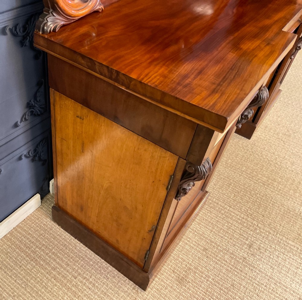 A Victorian mahogany pedestal sideboard featuring a carved arched backboard, flame mahogany veneer panels, and twin pedestal cupboards. View XV
