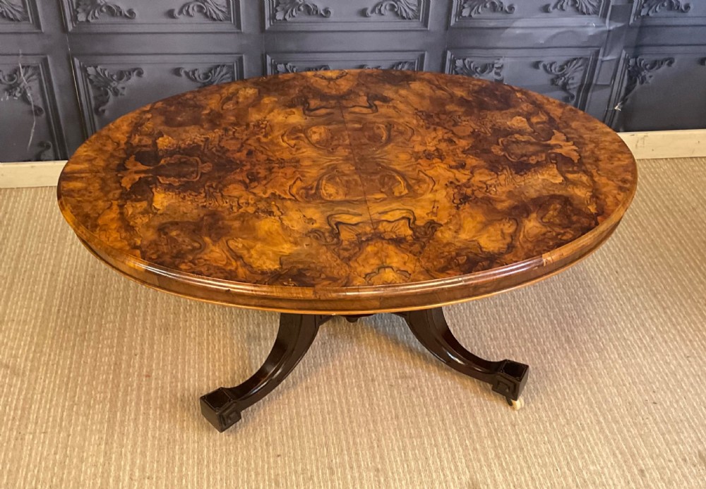 An oval Victorian low coffee table featuring a bookmatched burr walnut veneer top supported by dark curved legs on small castors.