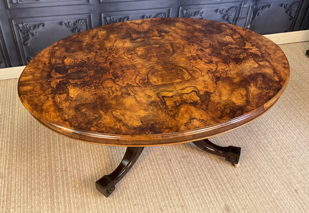 An oval Victorian low coffee table featuring a bookmatched burr walnut veneer top supported by dark curved legs on small castors. View VI