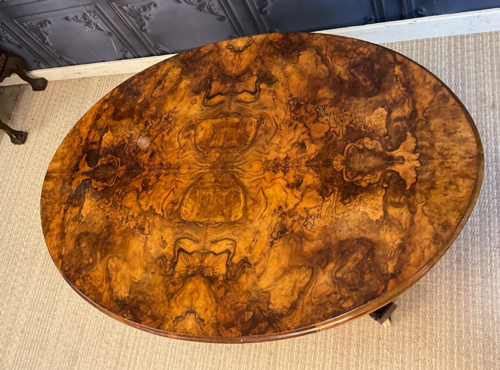 An oval Victorian low coffee table featuring a bookmatched burr walnut veneer top supported by dark curved legs on small castors. View IX
