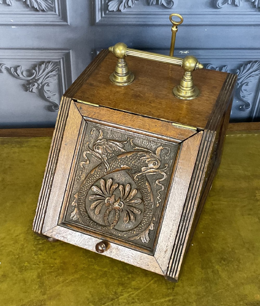 A Victorian carved oak coal scuttle featuring a brass top handle and a sloped front panel relief-carved with a mythical sea creature.