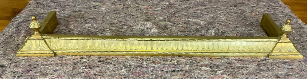 An antique Victorian brass fireplace fender featuring embossed leaf patterning along the curb and turned finials on each corner block. View II