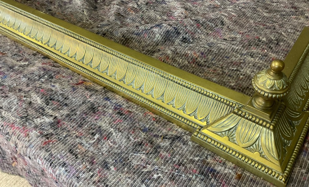 An antique Victorian brass fireplace fender featuring embossed leaf patterning along the curb and turned finials on each corner block. View V