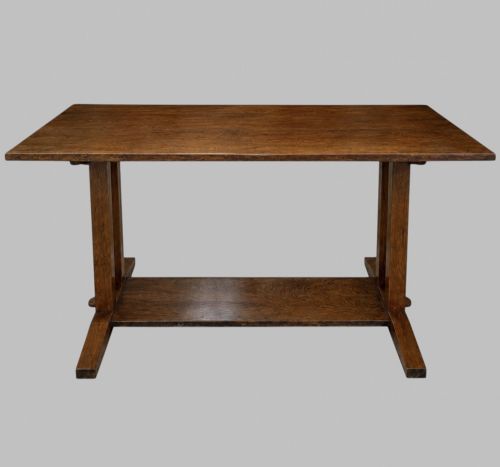 fig 1. An Arts and Crafts solid oak dining table featuring a rectangular top, twin upright trestle end supports, and a low solid wooden stretcher shelf.