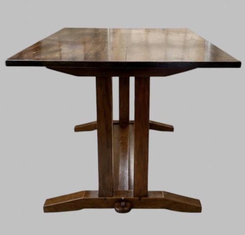 fig 2. An Arts and Crafts solid oak dining table featuring a rectangular top, twin upright trestle end supports, and a low solid wooden stretcher shelf.