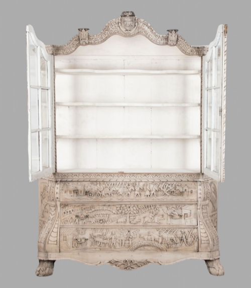 fig 1. A tall nineteenth-century Dutch carved pale oak display cabinet featuring a shaped crest, multi-pane glazed upper doors, and a bombe chest base with three relief-carved drawers on paw feet.