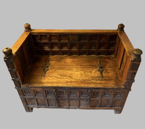 fig 1. A carved dark hardwood storage bench with a hinged lid seat, geometric panelled backrest, corner post finials, and iron fittings.