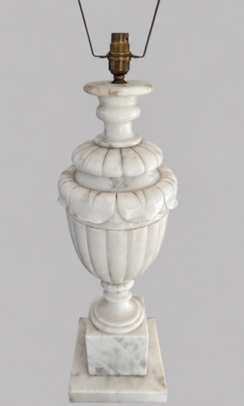 fig 1. A carved off-white alabaster table lamp base shaped like a classical urn with leaf motifs, resting on a stepped square plinth with a brass harp socket.