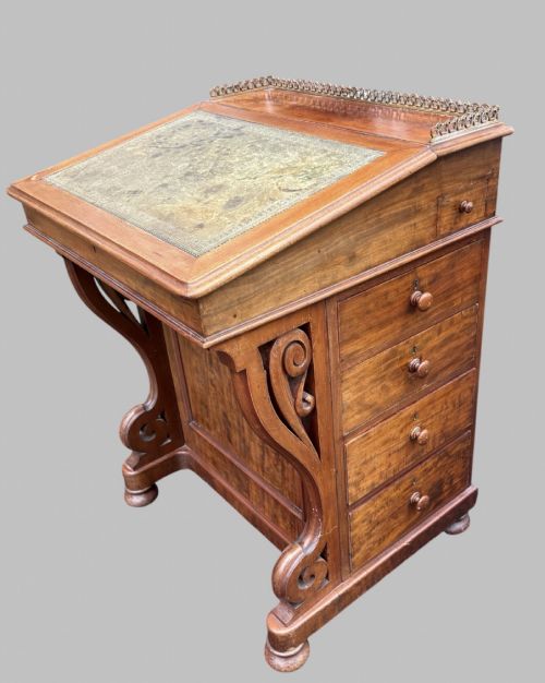 fig 1. A front view of a Victorian walnut Davenport desk featuring a slanted green leather writing surface, decorative top brass gallery rail, carved front supports, and a panelled wooden body.
