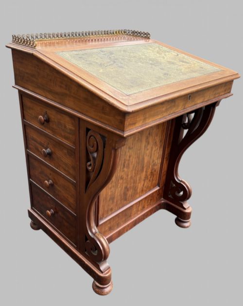 fig 2. A front view of a Victorian walnut Davenport desk featuring a slanted green leather writing surface, decorative top brass gallery rail, carved front supports, and a panelled wooden body.
