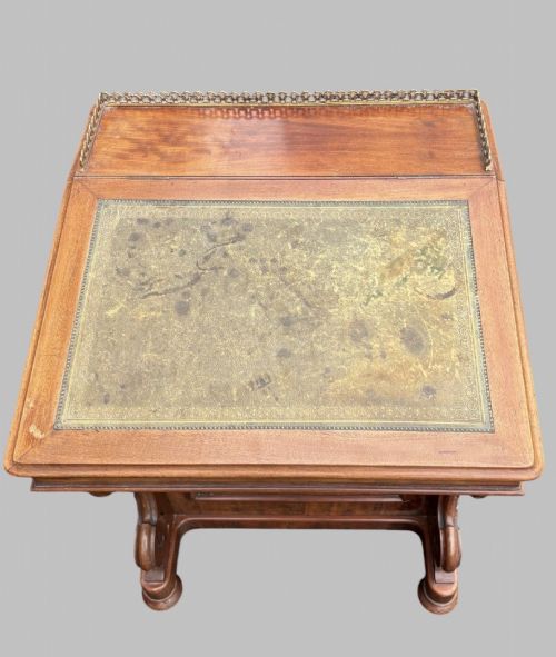 fig 3. A front view of a Victorian walnut Davenport desk featuring a slanted green leather writing surface, decorative top brass gallery rail, carved front supports, and a panelled wooden body.