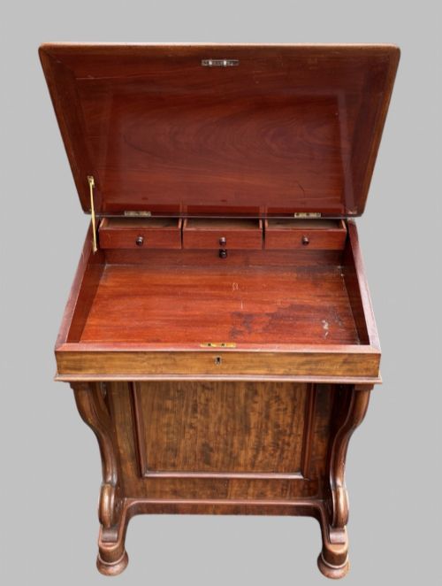 fig 4. A front view of a Victorian walnut Davenport desk featuring a slanted green leather writing surface, decorative top brass gallery rail, carved front supports, and a panelled wooden body.