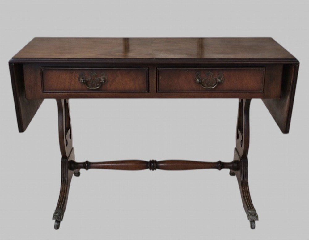 An early 20th-century mahogany drop-leaf table featuring two front drawers with brass swing handles, lyre-shaped end supports, a turned cross-stretcher, and splayed legs on brass castors.